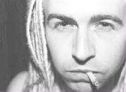 A close up, black-and-white photo of a man's face. He has a half-smoked cigarette in his mouth. He wears light coloured dreadlocks.