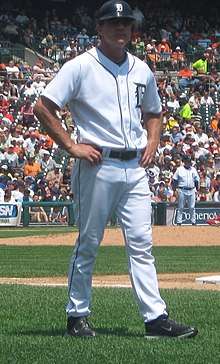 A man wearing a white baseball uniform and dark baseball helmet stands on a grass field with arms akimbo