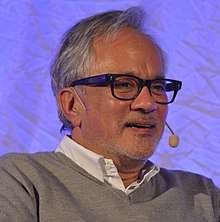 Photograph of an old man wearing black glasses and talking on mic.