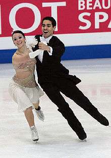 A man and woman ice dancing; the woman, on the left, is wearing a white and silver knee-length dress, and the man is wearing a classic tuxedo with a white shirt and bow-tie