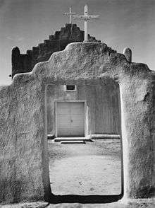 A black-and-white vertical photograph shows an adobe wall in the foreground, rising in the middle with a stairstep pattern and a white wooden cross at the pinnacle, with an open doorway beneath. Through the doorway and above the wall, an adobe church with white double doors and a similar stair-stepped roof and cross stands, slightly larger than the wall in front of it. The midday sun casts harsh shadows on the dirt ground.