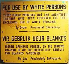 A rectangular yellow wall plaque. It contains a message written in black in both English and Afrikaans. In English, the message reads: "For Use by White Persons. These public premises and the amenities thereof have been reserved for the exclusive use of white persons. By Order Provincial Secretary".