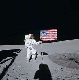 A astronaut in an Apollo space suit with red ring markings on the arms and legs and a red stripe down his helmet stands amid grey dust, holding a crumpled American flag on a flagpole