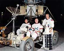 Three astronauts in space suits without helmets; two sit in a lunar rover mockup