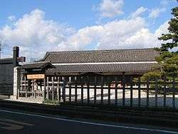 A wide wooden building with a large roof beyond a wooden fence.