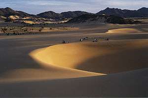 Sand dunes in the desert, offroad vehicles and mountains in the distance.