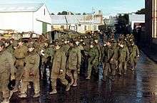 About forty soldiers wearing olive-green jackets and pants march down a wet urban street. They are guarded by three commandos with green berets and camouflage pattern jackets. In the background are paratroopers in similar camouflage jackets and maroon berets.