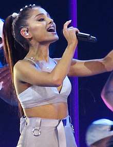 A woman with brown hair singing on stage wearing a lilac crop top and skirt