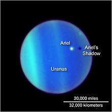 the planet Uranus is seen through the Hubble telescope, its atmosphere defined by bands of electric blue and green. Ariel appears as a white dot floating above it, casting a dark shadow below