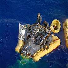 Photo of Armstrong and Scott in the Gemini capsule, in the water. They are being assisted by some recovery crew