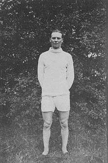 A man with dark hair is wearing a white top and white shorts. He is standing on a grass field.