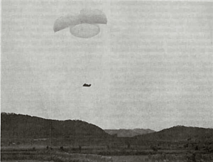 A dark shape is suspended in the air beneath three parachutes, while hills rise in the foreground