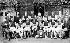 A group of men pose in two lines, one standing and the other seated. Eleven of the men are wearing dark-coloured football shirts with lighter sleeves, white shorts, shin pads and football boots. The remainder of the men are wearing formal suits. Almost all of the men sport moustaches. Displayed in the centre of the group are two large trophies.