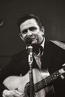 A dark-haired man wearing a black jacket, playing a guitar and singing into a microphone