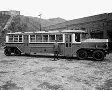 A long, 8-wheeled city bus is viewed from the side, parked in front of a brick building. The bus is about 1.5 times as tall as the uniformed man in the foreground.