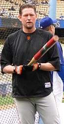 A man in a black baseball uniform pointing a bat towards the camers