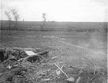 Two soldiers operate a field gun from a dug-in position in an open field and reinforced with sandbags. Entrenching tools, spoil and broken vegetation lie around the emplacement, while a number of defoiliated trees stand in the middle distance.