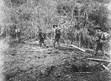 Soldiers patrol through a jungle setting, passing a makeshift grave marker made out of bamboo