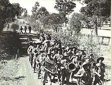 Soldiers marching along a dirt road in the Australian bush, led by a brass band