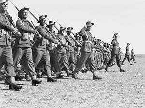 Black and white photo of a large group of men wearing military uniforms and carrying rifles marching in close formation