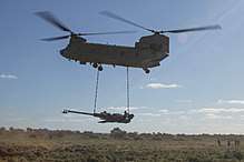 Colour photo of a twin rotor helicopter flying near the ground with an artillery gun slung below it