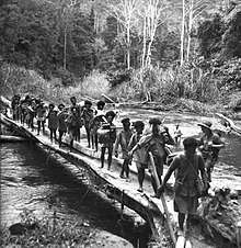 Black and white photo of Melonesian men crossing a log bridge across a river while carrying loads. A Caucasian man wearing military uniform is standing on the bridge, and two other Caucasian men are swimming in the river.