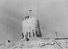 Soldiers stand at the base of a sandstone structure in the desert whilst a flag is unfurled on top of the building