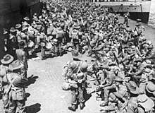 Black and white photo of a large number of men wearing military uniforms on a dock. Most of the mean are wearing brimmed hats and are carrying rifles.