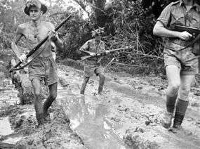A black and white photograph of Australian soldiers patrolling along a muddy track surrounded by thick jungle. The men are armed with rifles which are held at the high port position and are wearing tropical uniforms consisting of shorts and lightweight short sleeve shirts. One of the soldiers is patrolling without a shirt.