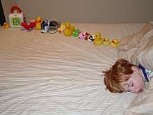 Young boy asleep on a bed, facing the camera. On the bed behind the boy's head is a dozen or so toys carefully arranged in a line.