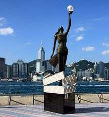 A bronze statue on a pedestal, with the Hong Kong skyline in the background. The pedestal is designed in the image of four clapperboards forming a box. The statue is of a woman wrapped in photographic film, looking straight up, with her left hand stretched upwards and holding a glass sphere containing a light.