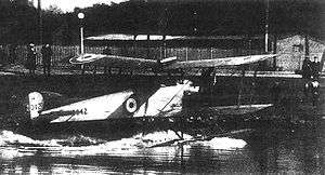 Three-quarter rear view of biplane on floats, taxiing along a stretch of water towards shore