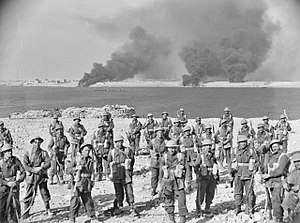 A group of soldiers stand on a foreshore. Smoke billows in the background.
