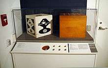 A museum exhibit comprising a table, in the corner of a room between a door with a fire alarm handle, with a glass case on top containing two wooden boxes with holes in the top. The front of the table comprises a printed description of the exhibits within the case and several holes, simulating the artworks within the case.