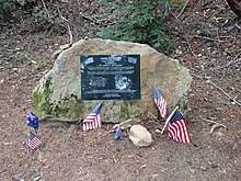 Photo of a memorial plaque fixed to a rock in El Corte de Madera preserve. Multiple small flags of the United States and Australia have been placed in front of it.