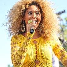 With curly brown hair, a woman holds a microphone looking down in front of her.