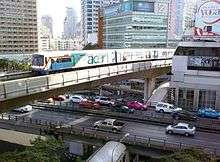 An elevated train, painted in blue, white and a red stripe and with advertisements with the name &quot;acer&quot;, running above a road lined with many tall buildings and crossing an intersection with a flyover bridge with many cars