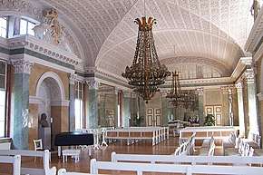Photograph of a Baroque interior of a hall in Palace, with stucco ceiling and a large chandelier hanging from it, all in light colours