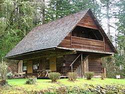 Photograph of a two-story log cabin, with an extreme overhang of the second story over the first