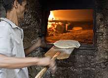A man using a bread peel to slide a round disk of raw flatbread dough into a brick oven