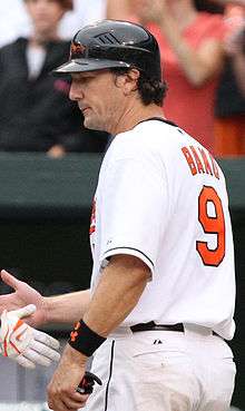 A man in a white baseball jersey with orange lettering reading "BAKO" and "9" and a black baseball helmet shakes hands with an unseen person