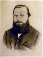 A man in his late 20s or early 30s with dark hair and a bushy beard, wearing a dark coat, dress shirt and tie.