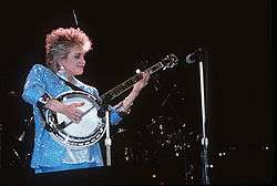 A blonde-haired woman wearing a sparkly blue jacket playing a banjo on stage