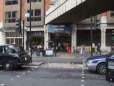Across a road with a London taxi and a car is an entrance. This has people standing in it and above is a blue rectangular sign reading "BARBICAN STATION" in white and above this is a bridge linking the building