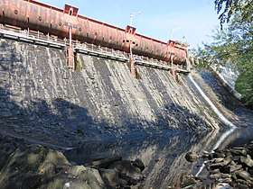 A high dam of irregular square black stone. The top of the dam is an orange primered spillway held in position by hydraulic jacks built into notches in the dam face. A spillway runs down both sides of the dam into a small rocky pond at the foot of the dam. The dam is in the forest and some trees intrude into the picture.