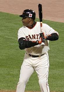A man in a white baseball uniform stands in the left-handed batter's box. He is holding a black bat and wearing a black batting helmet. His uniform reads "Giants" across the chest.