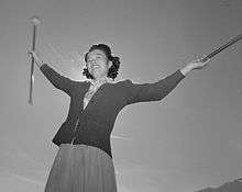 Japanese teenage girl in 1940s sweater, skirt, and blouse twirling two batons and smiling, backlit by the sun against a nearly-cloudless sky.