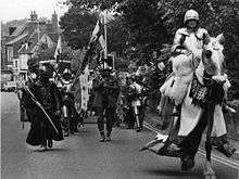 An armoured and mounted man leads a small party, similarly dressed in mediaeval attire, along a road.