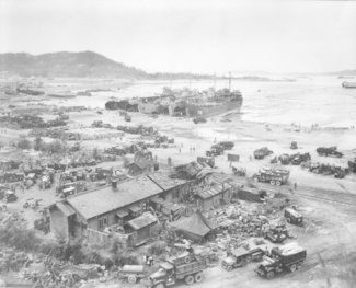 A village by a beach filled with landing craft, vehicles, and troops from a recently landed force
