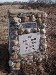 Stone cairn erected in 1975 marking the Battleford Industrial School Cemetery. A plaque at the top of the cairn reads: RESTORATION THROUGH OPPORTUNITIES FOR YOUTH, 4S1179-1974. PLAQUE PROVIDED BY DEPARTMENT OF TOURISM AND RENEWABLE RESOURCES.